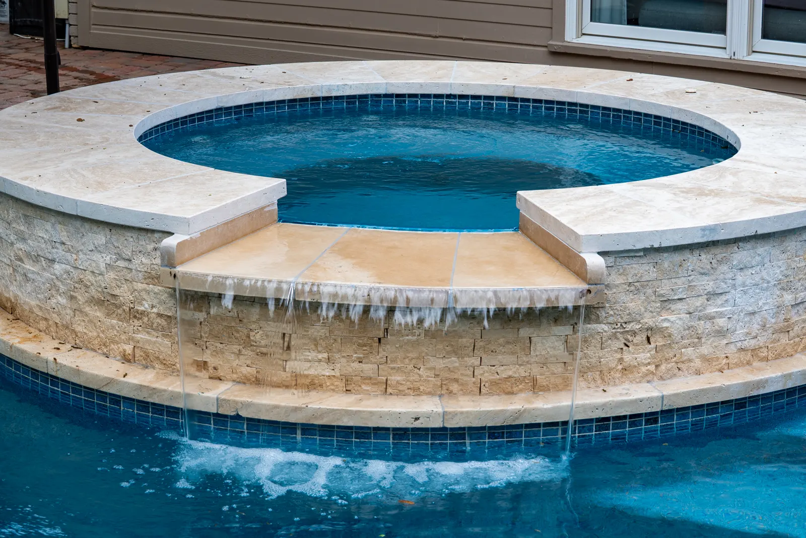 Round raised spa with travertine coping spilling into a freeform pool