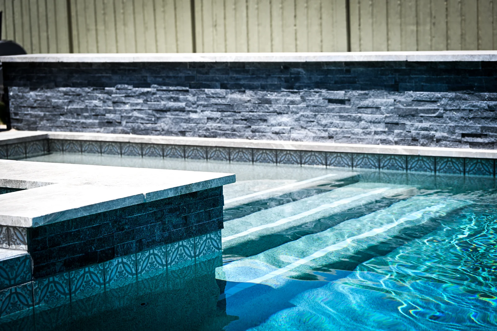 Dark stacked-stone wall above patterned waterline tile, with sunlit pool steps
