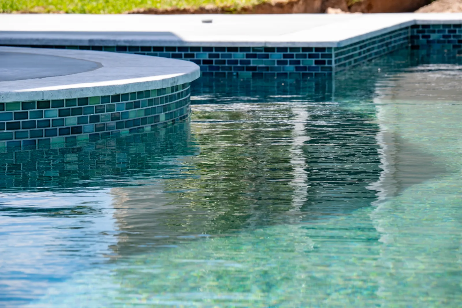 Edge of a raised spa faced in teal glass tile, reflected in the pool