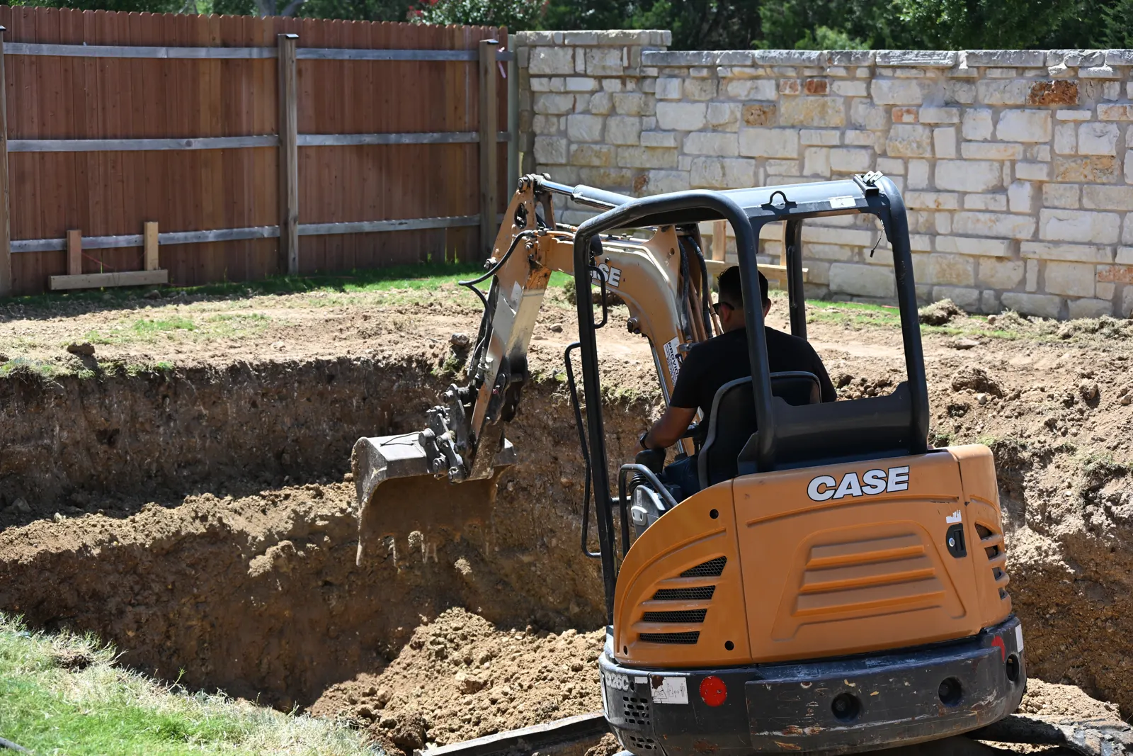 Mini excavator cutting the pool shape into a lawn beside a stone wall